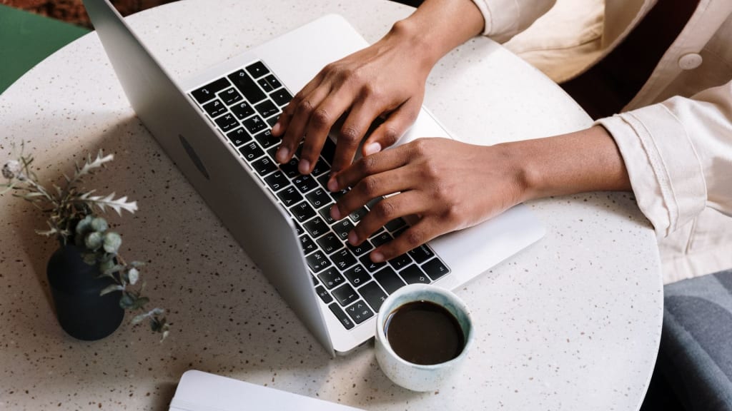 A person typing on a laptop at a round table with a small plant and a cup of coffee nearby.